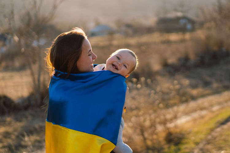 Woman hugs her little son wrapped in yellow and blue flag of ukraine in outdoors.