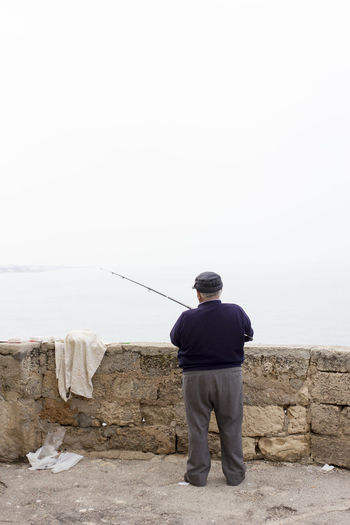 Rear view of senior man fishing while standing by retaining wall at beach against sky