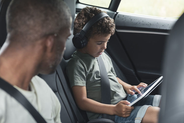 Man looking at son watching video on digital tablet in electric car