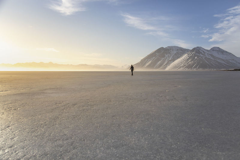 Distant anonymous person walking alone through plain desert terrain covered with white snow under cloudless sky in winter day