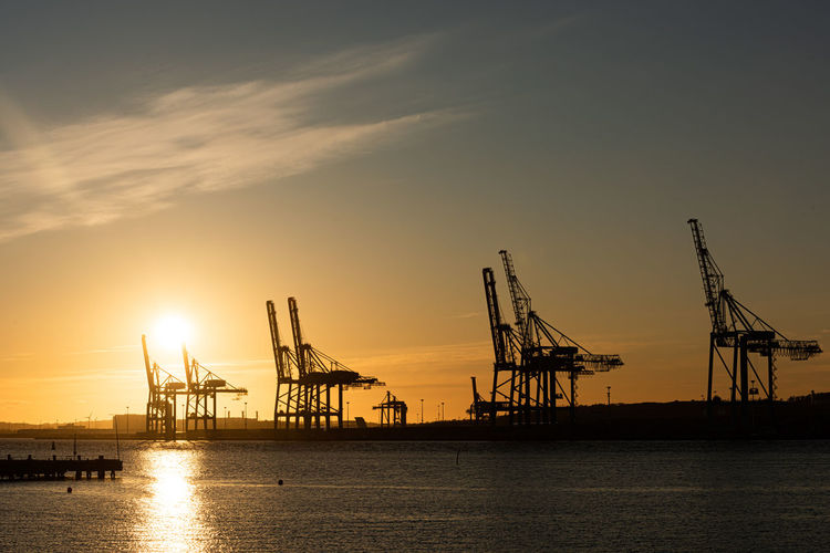 Silhouette cranes at commercial dock against sky during sunset