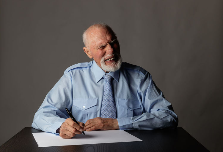 Senior businessman writing in paper and making face while sitting on table against gray background