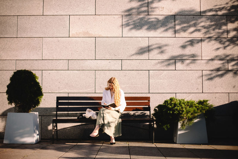 Young woman writing in diary sitting on bench by wall in summer city