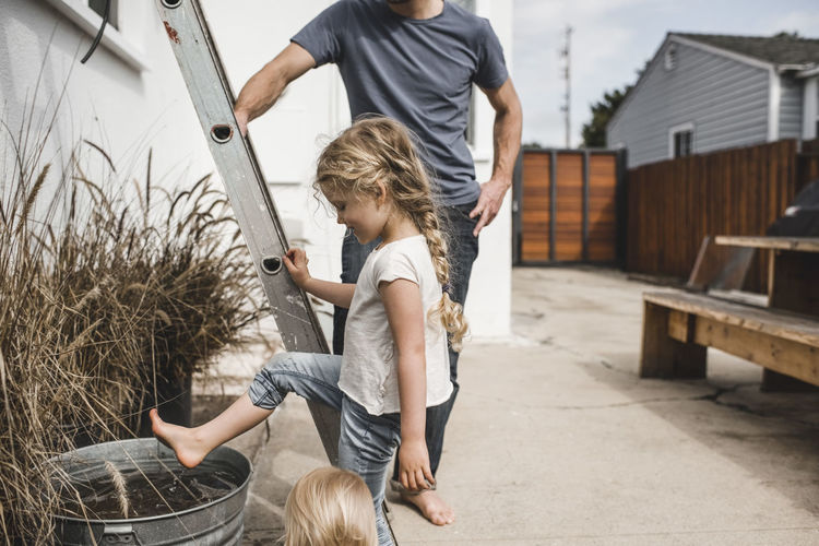 Girl playing with water in container while standing by father and ladder outside house