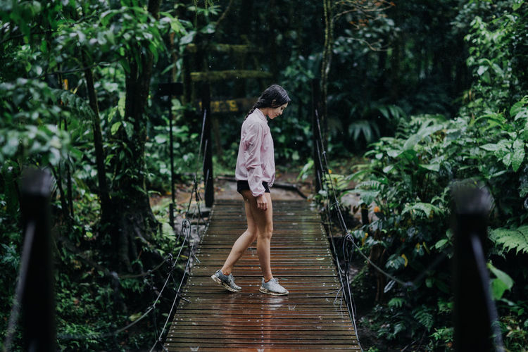 Full body side view of young female traveler standing on narrow suspension bridge over celeste river with lush green tropical vegetation during summer adventure in costa rica