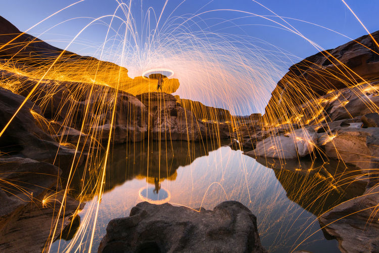 Light trails over lake on rock against sky at dusk
