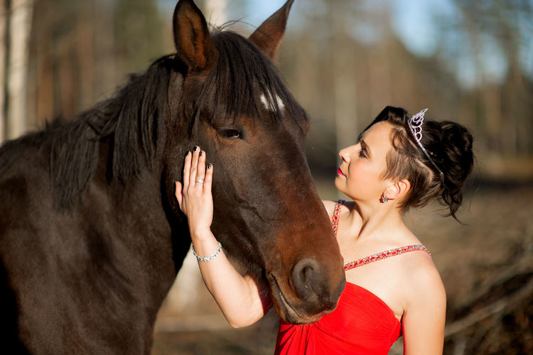 Beautiful woman wearing a red dress posing with a brown horse outdoor