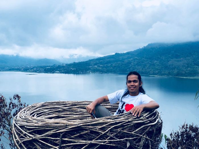 Portrait of man sitting in large nest with lake in background against cloudy sky