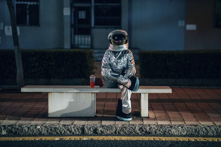 Spaceman sitting on bench at a bus stop at night with soft drink