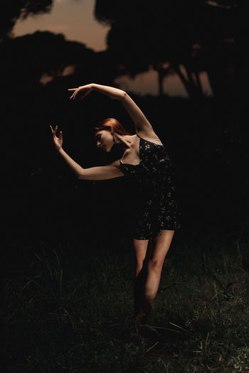Elegant female ballet dancer in dress standing in meadow with raised arms and performing in dark park in iceland