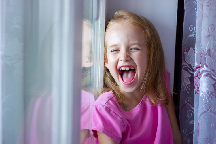 Little girl in a pink dress sits on the windowsill and laughs with her mouth open, happy childhood