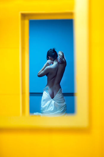 Rear view of bride getting dressed seen through yellow window