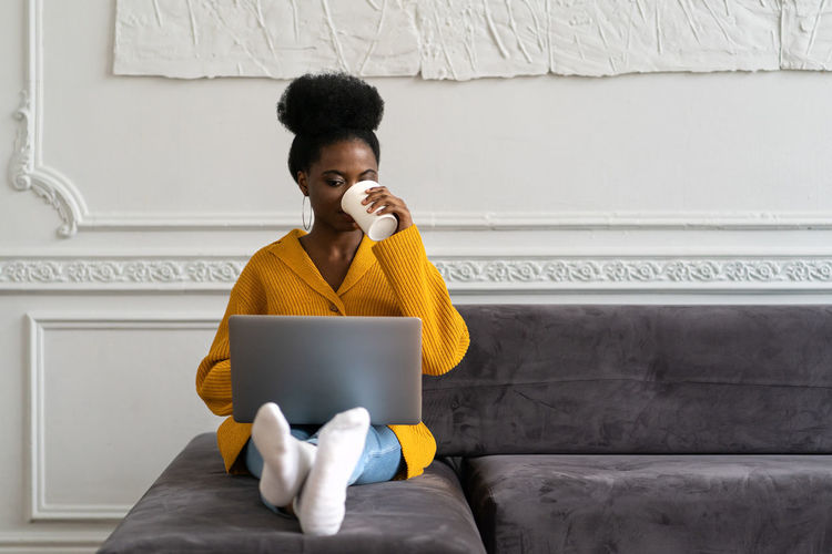 Afro-american woman sitting on couch, working online on laptop, watching webinar or video, drink tea