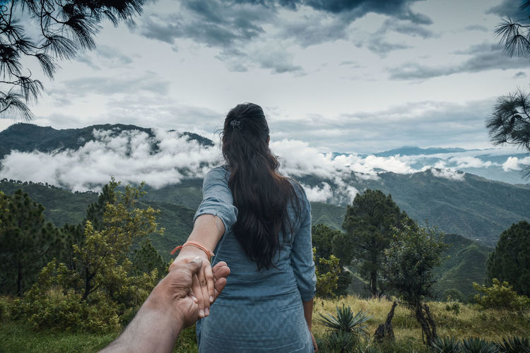 Cropped image of man holding woman hand on mountain against cloudy sky