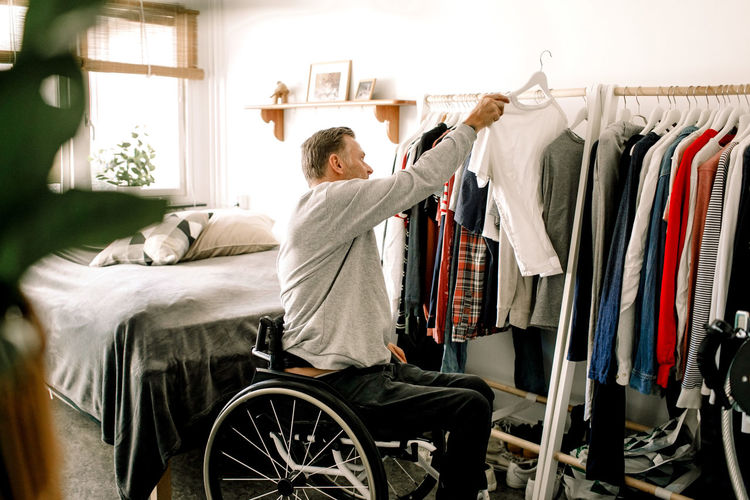 Side view of disabled mature man hanging white t-shirt on clothes rack at home