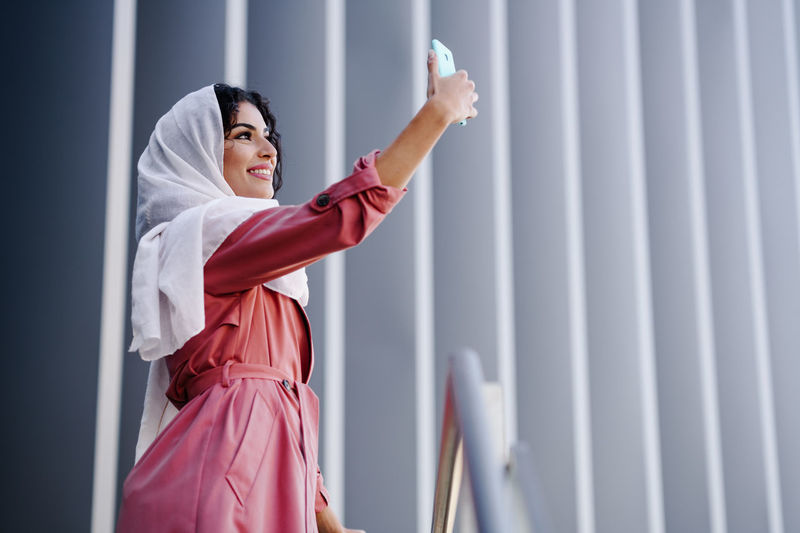 Woman taking selfie while standing against wall