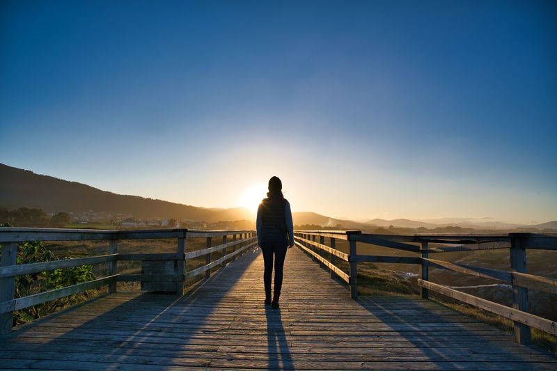 Rear view of woman standing on boardwalk against sky during sunset