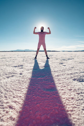 Full length of man flexing muscles while standing on sand at beach against sky during sunny day