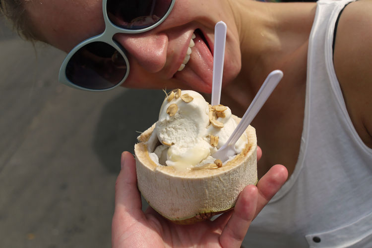 Woman licking coconut ice cream held by cropped hand on sunny day