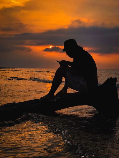 Silhouette man using mobile phone while sitting on driftwood at beach against sky during sunset