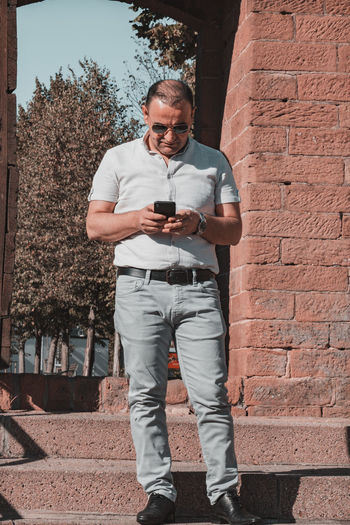 Full length of young man using phone while standing against brick wall