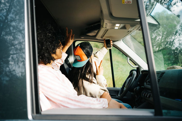 Through window side view of cheerful young female friends having fun and taking selfie on smartphone while sitting inside camper vehicle during summer trip in nature