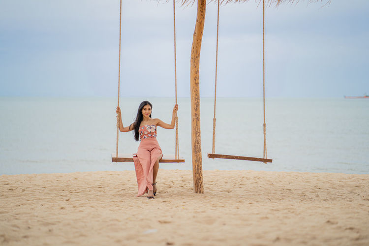 Full length of woman sitting on rope swing at beach against sky