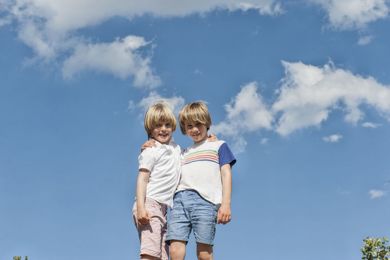 Low angle of adorable brothers hugging against blue sky in summer while looking at camera and enjoying weekend