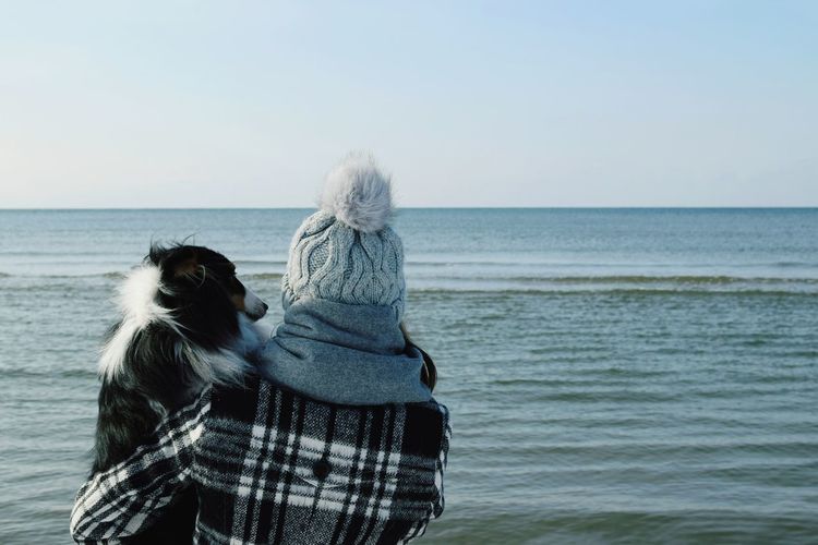 Woman in warm clothing holding dog against sea