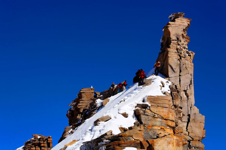 Low angle view of people on rocky mountain during winter against clear blue sky