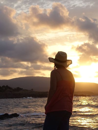 Rear view of woman wearing hat standing by lake against sky during sunset