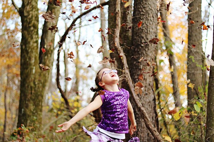 Girl throwing autumn leaves by bare trees in forest