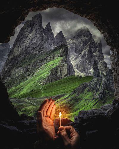 Cropped hand holding burning matchstick in cave against mountains