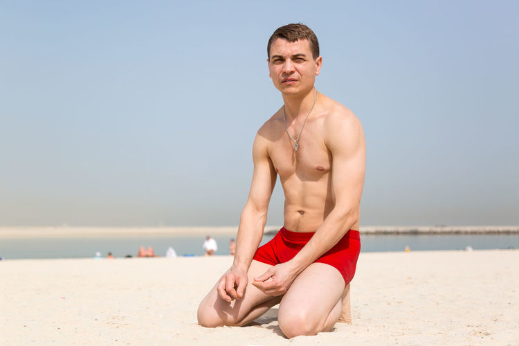 Portrait of young man sitting on beach against clear sky