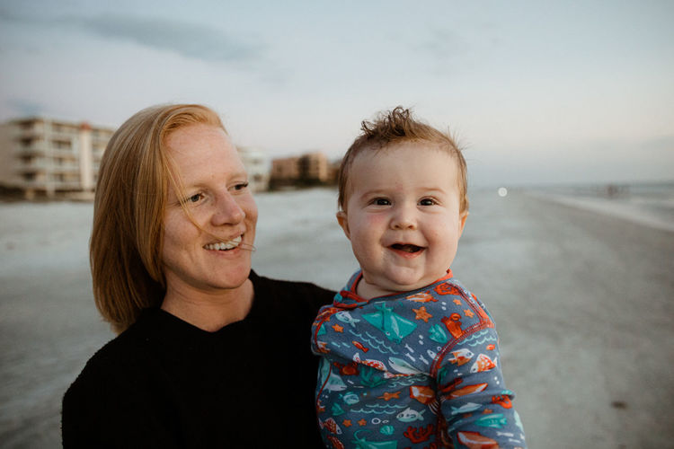 Fat baby boy with crooked smile held by young red haired mom on beach