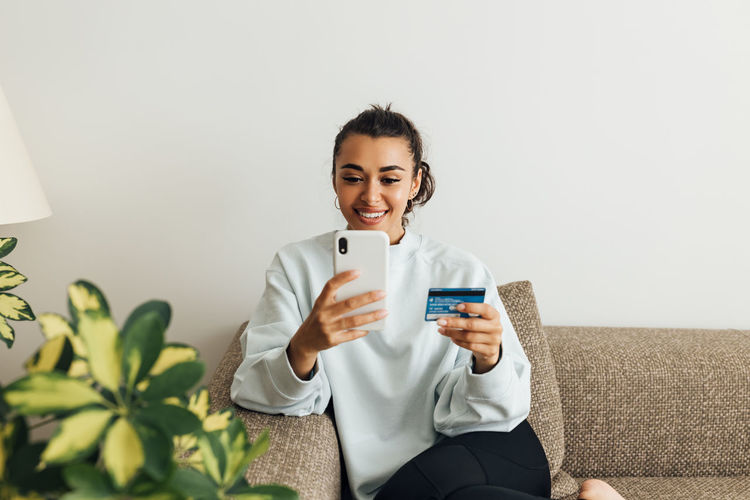 Smiling woman holding credit card while using smart phone at home