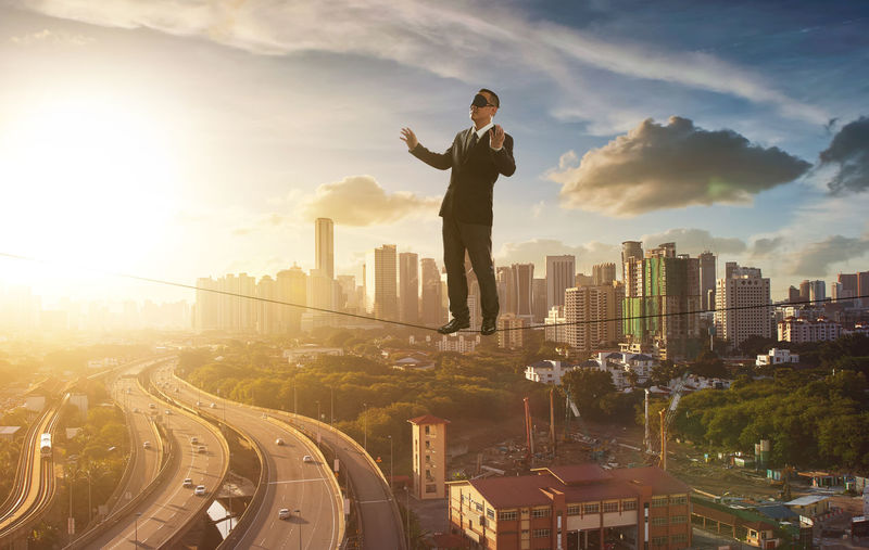 Full length of businessman with blindfold standing on rope in city during sunset