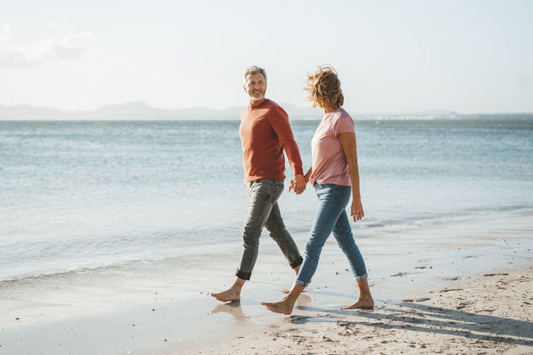 Happy mature couple holding hands walking on shore at beach