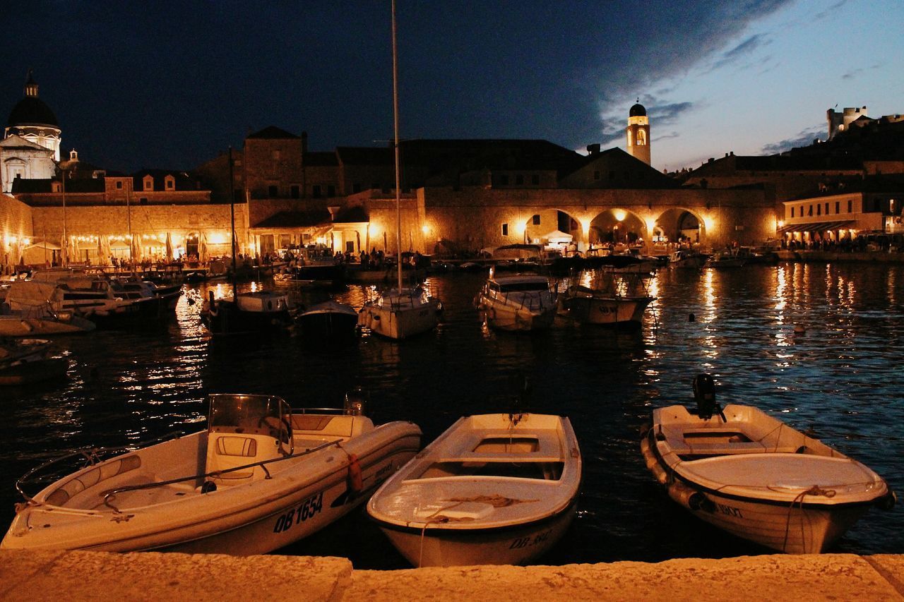 BOATS MOORED ON RIVER BY ILLUMINATED CITY AT NIGHT