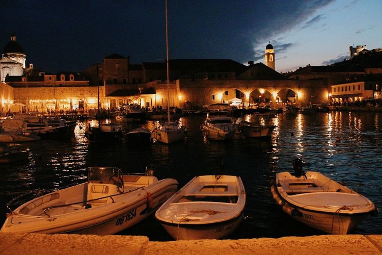 Boats moored on river by illuminated city against sky at night