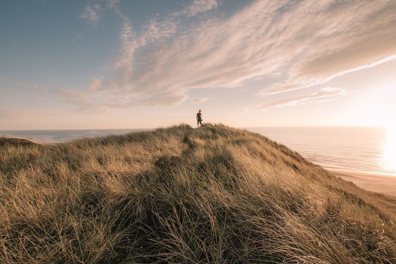 Rear view of man standing on mountain by sea against sky during sunset