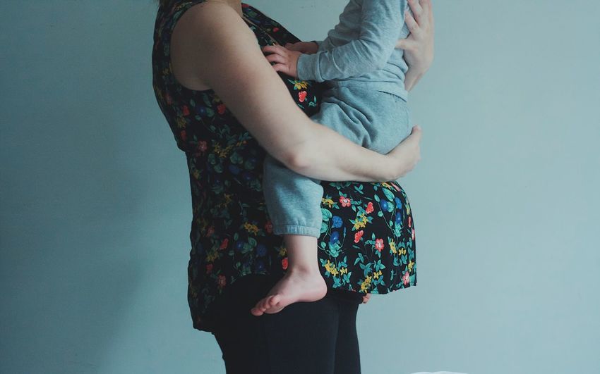 Cropped image of pregnant woman carrying son while standing against wall