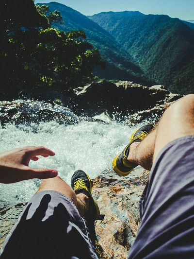 Low section of man sitting by waterfall against mountains