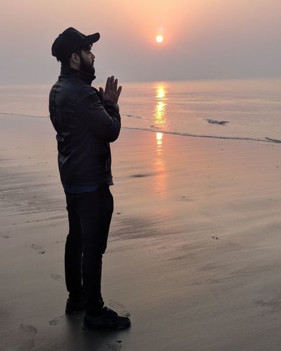 Young man praying while standing at beach during sunset
