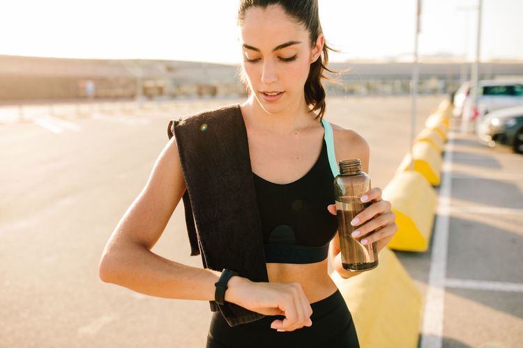 Young fit female in sportswear holding bottle of water and checking sports results on activity tracker while resting after running on street