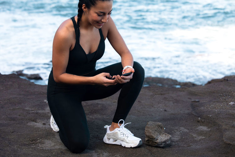 High angle side view of athletic female in activewear leaning on knee on beach near sea and surfing internet on smartphone