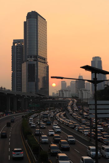 Traffic on road by buildings against sky during sunset