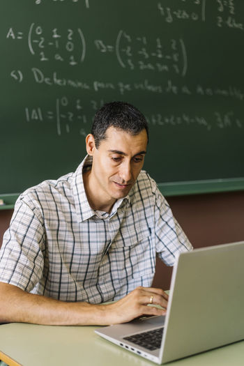 Young man using laptop while sitting on blackboard