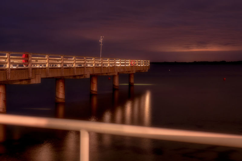 Illuminated pier over river against sky at sunset