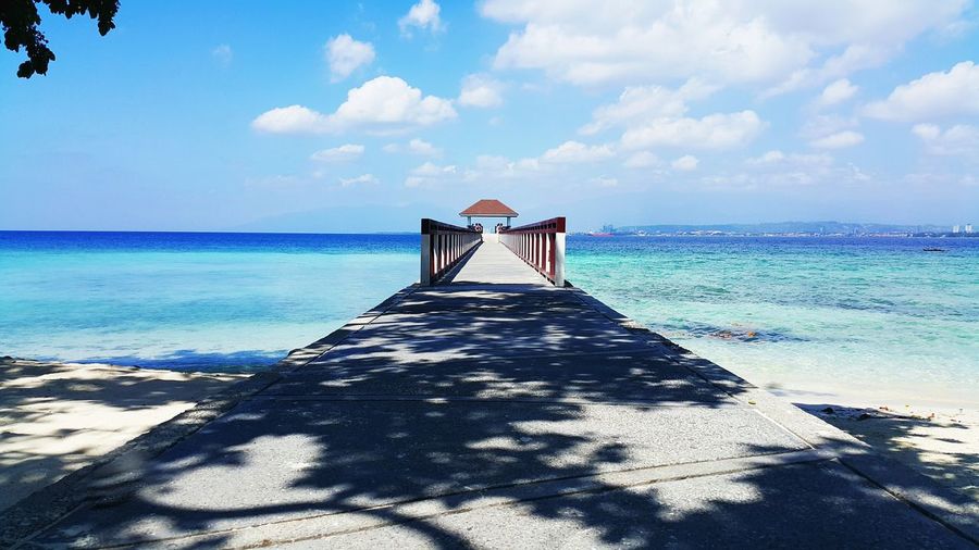 Pier leading towards gazebo at beach against cloudy sky during sunny day